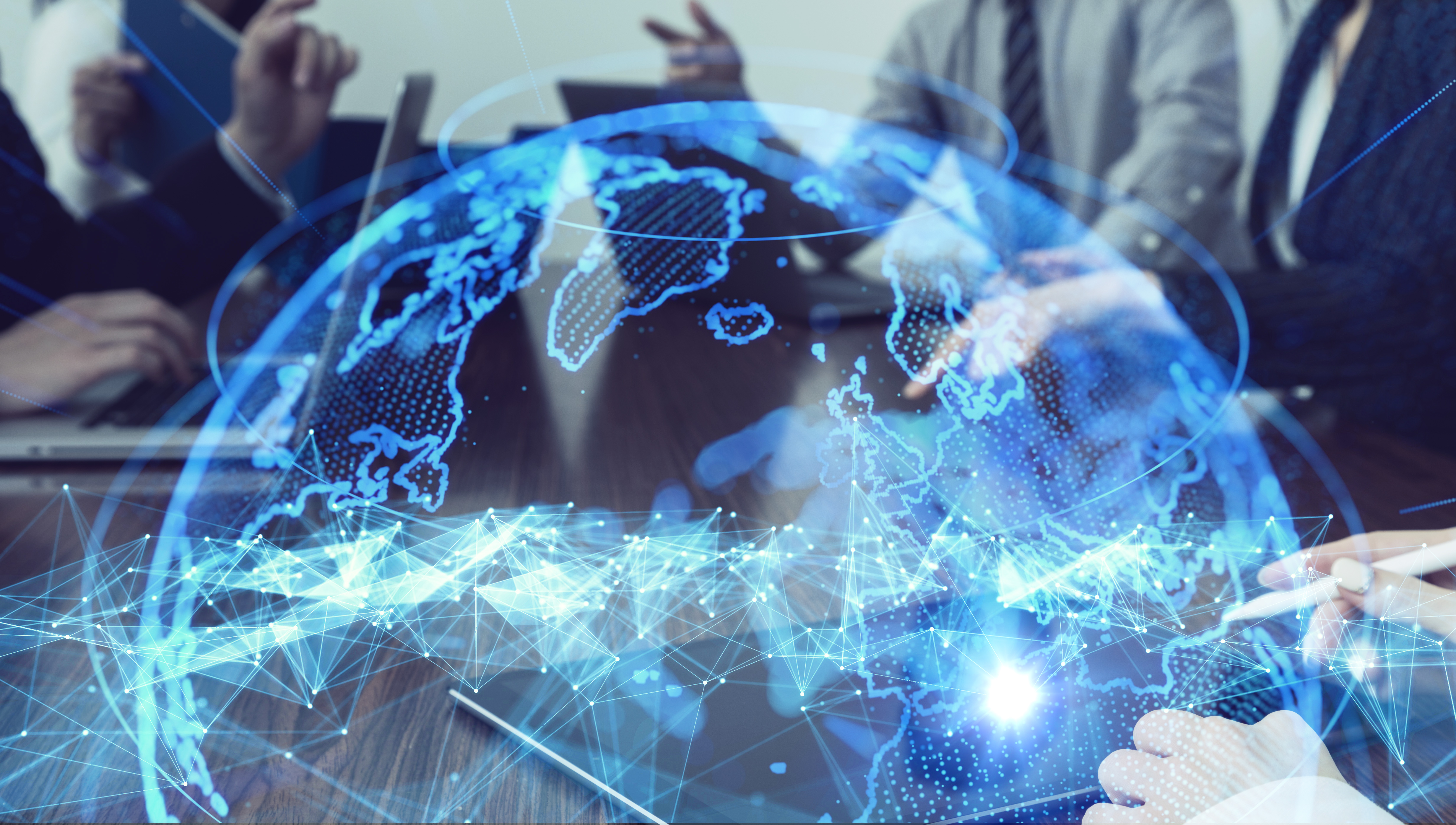 Eine digitale Weltkarte mit leuchtenden Linien und Punkten, die Vernetzung symbolisieren. Im Hintergrund sind Hände zu sehen, die an Laptops arbeiten, während sich die Personen in einer Besprechung befinden. Die Darstellung vermittelt das Gefühl von globaler Kommunikation und Zusammenarbeit.