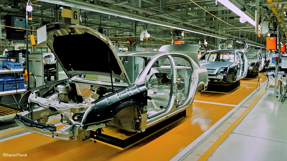 The image shows an automotive assembly line with several car chassis in various stages of production. The foreground features an open chassis, while others are lined up in the background, indicating a busy manufacturing environment.