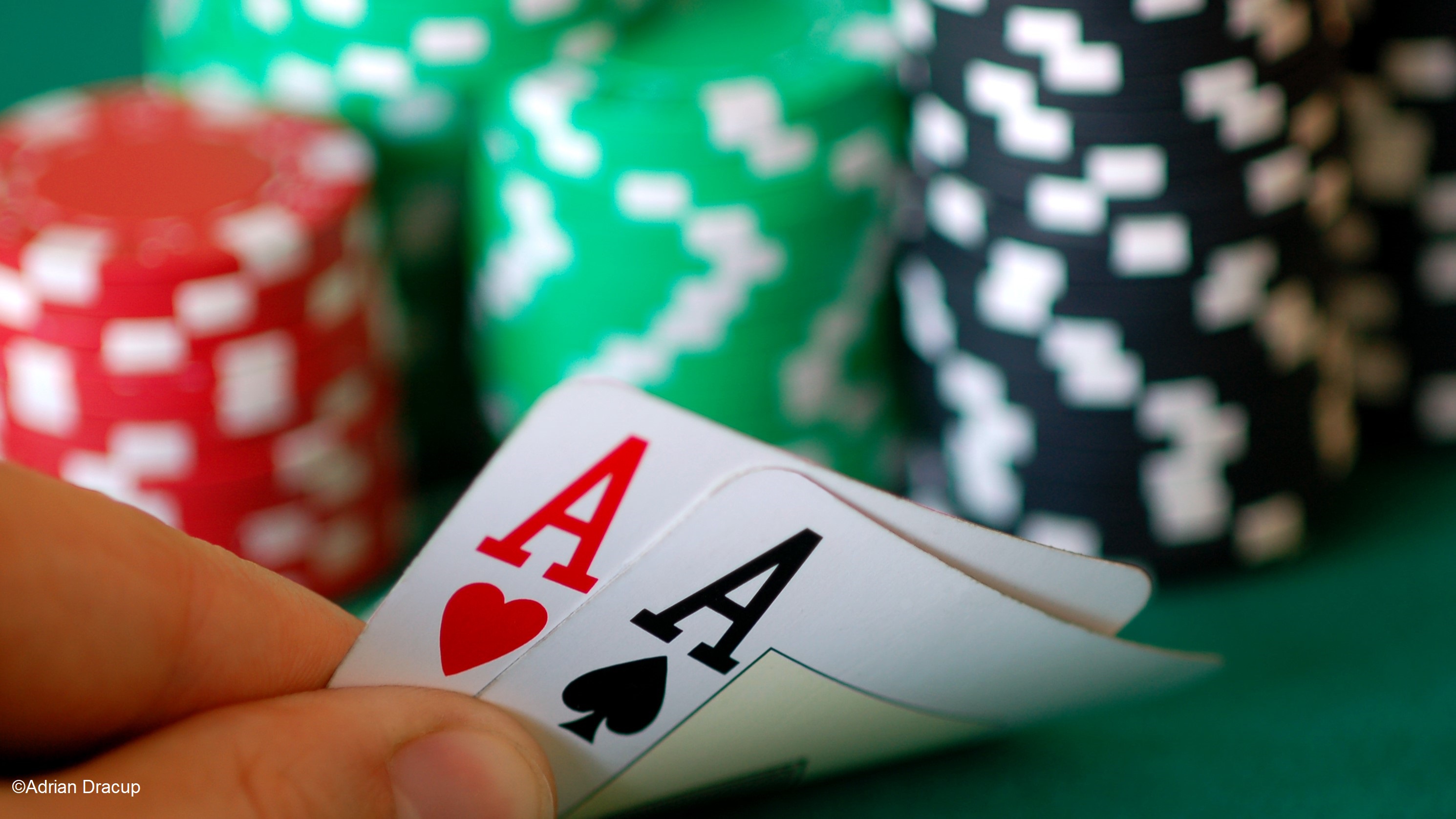 A close-up of a hand holding poker cards showing two Aces, one red and one black, while colorful poker chips in red, green, and black are stacked in the background. The setting appears to be a poker table.