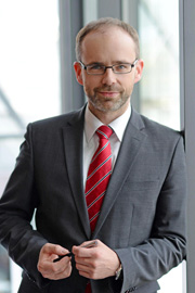 A man in a gray suit stands in front of a large window, holding a pen in his hands. He has short hair and wears glasses, with a red tie adding color to his business attire. The background is bright and modern.