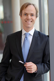 A man in a dark suit and light blue tie stands confidently next to a window, holding a pen in his right hand. He has short, light brown hair and is smiling, exuding a professional and approachable demeanor.