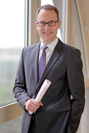Ein lächelnder Mann in einem Anzug steht vor einem Fenster. Er trägt eine Brille und hält ein Dokument in der Hand. Der Hintergrund ist hell und dezent.
