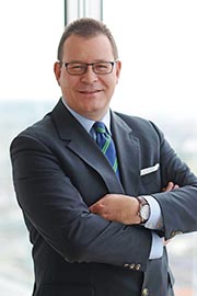 Ein Mann im Anzug mit Brille steht mit verschränkten Armen vor einem großen Fenster, das einen Blick auf die Stadt bietet. Er lächelt freundlich und trägt ein gestreiftes Hemd sowie eine Krawatte in Blau und Grün.