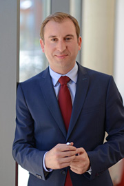 Porträt eines Mannes in einem dunkelblauen Anzug mit roter Krawatte. Er steht lächelnd an einem Fenster und hält einen Stift in der Hand. Der Hintergrund ist hell und modern eingerichtet.