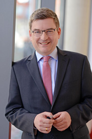 Ein lächelnder Mann trägt einen Anzug mit Krawatte und steht an einem Fenster. Er hält einen Stift in der Hand und wirkt freundlich und selbstbewusst. Das Bild vermittelt einen professionellen Eindruck.