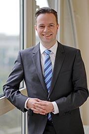 Ein lächelnder Mann in einem Anzug steht neben einem Fenster. Er trägt ein hellblaues Hemd mit einer gestreiften Krawatte und hat die Hände vor sich verschränkt. Der Hintergrund ist unscharf, deutet aber auf ein Büro- oder Geschäftsumfeld hin.