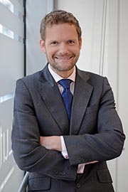 Ein lächelnder Mann in einem eleganten Anzug steht mit verschränkten Armen. Er hat kurze, blonde Haare und trägt eine Krawatte. Der Hintergrund ist hell und modern gestaltet.