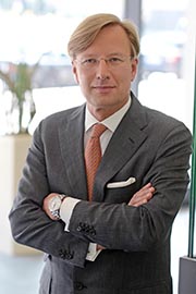 Ein Mann in einem grauen Anzug steht mit verschränkten Armen vor einer modernen Umgebung. Er trägt ein weißes Hemd und eine orangefarbene Krawatte. Der Ausdruck ist selbstbewusst und freundlich.