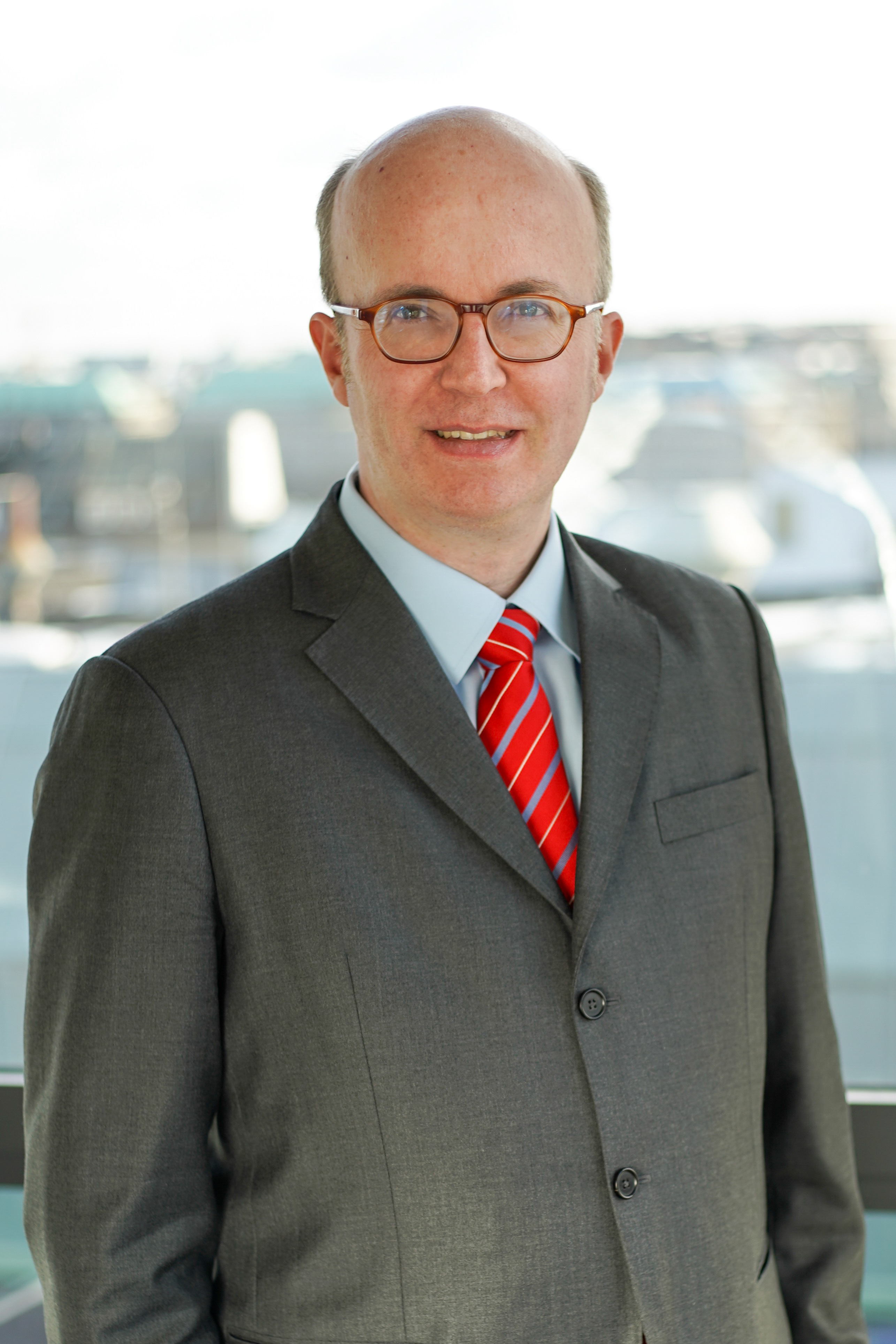 Ein Mann steht vor einem großen Fenster, durch das Tageslicht strömt. Er trägt einen dunklen Anzug mit einem rot-gestreiften Schlips und hat eine Brille auf. Der Hintergrund zeigt unscharfe Gebäude.