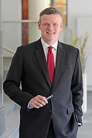 Ein lächelnder Mann in einem schwarzen Anzug und roten Krawatte steht mit einem Stift in der Hand. Der Hintergrund zeigt eine moderne Büroumgebung.