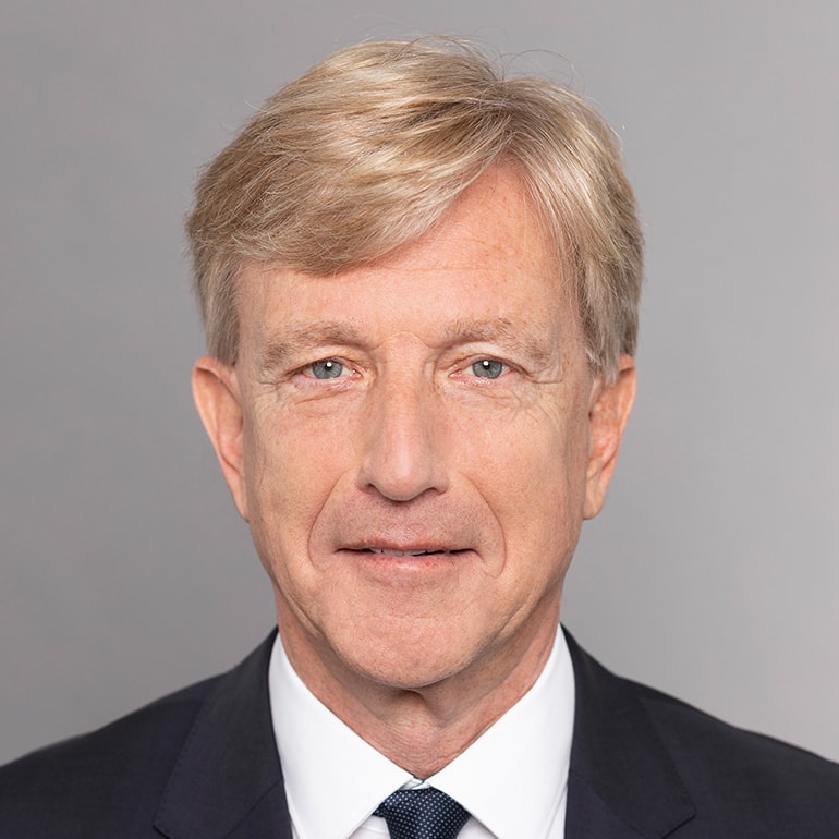 Ein älterer Mann mit blonden Haaren und einem freundlichen Gesichtsausdruck trägt einen dunklen Anzug mit einer hellen Krawatte. Er schaut direkt in die Kamera vor einem neutralen grauen Hintergrund.