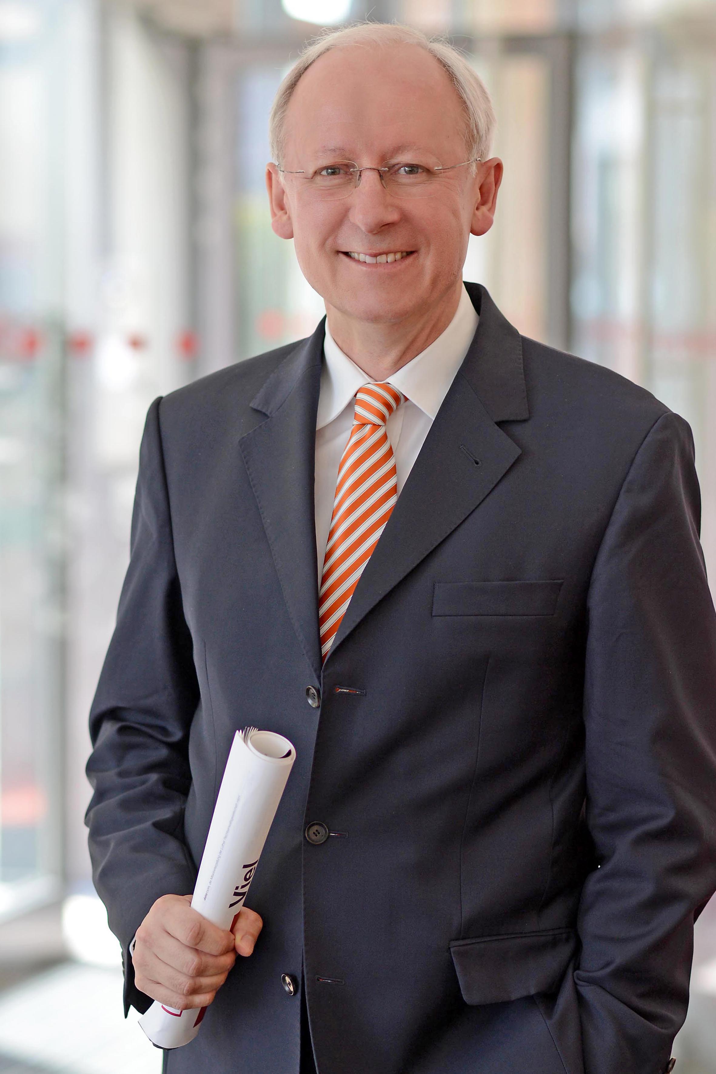 Ein Mann in einem dunklen Anzug mit einer gestreiften orange-weißen Krawatte lächelt freundlich. Er hält eine gerollte Dokumentenmappe in der Hand und steht vor einem unscharfen Hintergrund.