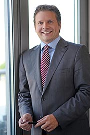 A man in a business suit stands near a window, smiling confidently. He holds a pen in one hand and appears approachable and professional, suggesting a business or corporate setting.