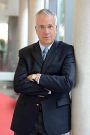 Ein Mann in einem dunklen Anzug steht mit verschränkten Armen an einer Säule. Er hat kurze, graue Haare und trägt ein helles Hemd mit einer Krawatte. Der Hintergrund ist unscharf, aber hell erleuchtet.