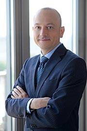 Ein Mann in einem dunkelblauen Anzug mit Krawatte steht an einem Fenster und lächelt. Er hat die Arme vor der Brust verschränkt und strahlt Selbstbewusstsein aus.