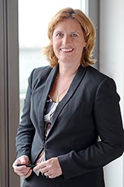 Eine lächelnde Frau in einem schwarzen Blazer steht an einem Fenster. Sie hält einen Stift in der Hand und wirkt selbstbewusst. Der Hintergrund ist hell und modern.