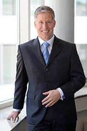 Ein lächelnder Mann in einem dunklen Anzug steht neben einem Fenster. Er trägt ein hellblaues Hemd und eine gestreifte Krawatte. Die Position vermittelt Selbstbewusstsein und Professionalität.