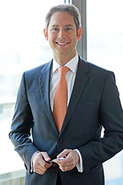 Ein lächelnder Mann in einem dunklen Anzug mit einem hellen Hemd und einer orangefarbenen Krawatte steht an einem Fenster. Er hält eine Brille in der Hand und wirkt freundlich und professionell.