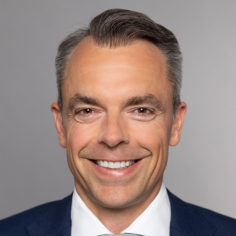 Ein lächelnder Mann mit kurzen, grauen Haaren trägt einen dunkelblauen Anzug und eine helle Krawatte. Der Hintergrund ist hellgrau. Der Ausdruck des Mannes wirkt freundlich und einladend.