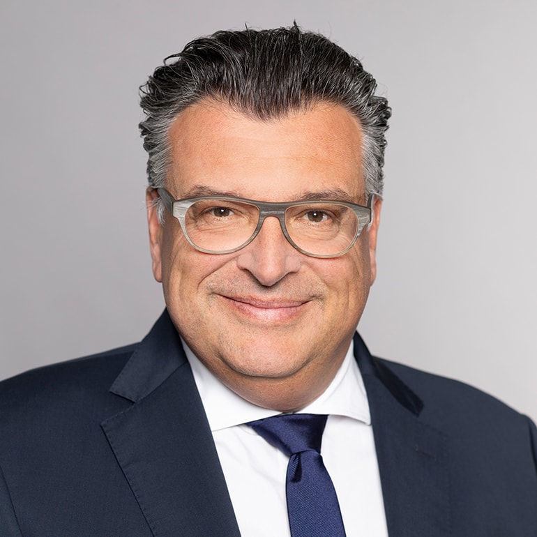 Porträt eines lächelnden Mannes in einem dunklen Anzug mit einem weißen Hemd und einer blauen Krawatte. Er trägt eine modische Brille und hat mittellange, graue Haare. Der Hintergrund ist neutral grau.