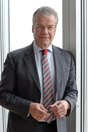 Ein Mann steht an einem Fenster und trägt einen Anzug mit einer gestreiften Krawatte. Er hält einen Stift in der Hand und lächelt leicht. Der Hintergrund lässt Licht herein und betont die seriöse Atmosphäre.