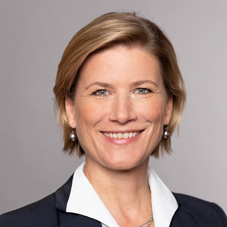 Porträt einer lächelnden Frau mit kurzem, blondem Haar, die einen blauen Blazer und ein weißes Hemd trägt. Der Hintergrund ist einfarbig und neutral. Die Frau sieht freundlich und selbstbewusst aus.