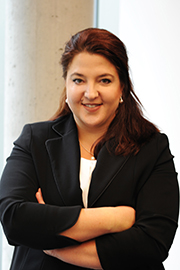 Eine professionell gekleidete Frau mit langen, braunen Haaren steht lächelnd vor einer hellen Wand. Sie trägt einen schwarzen Blazer und hat die Arme vor der Brust verschränkt. Die Bildkomposition vermittelt Selbstbewusstsein und Professionalität.