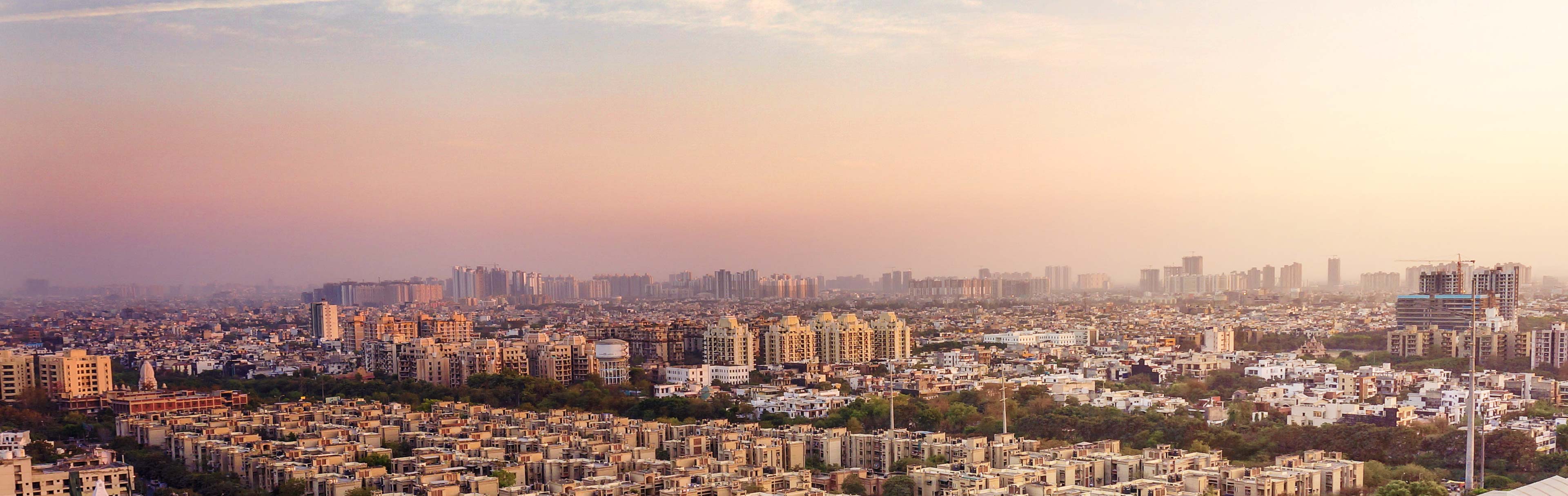 Blick auf eine weitläufige Stadtlandschaft mit vielen Wohngebäuden, die sich bis zum Horizont erstreckt. Die Farben des Himmels variieren von sanften Lila- und Rosatönen, was auf einen Sonnenuntergang hindeutet. Die Szene vermittelt ein Gefühl von Weite und städtischer Dichte.