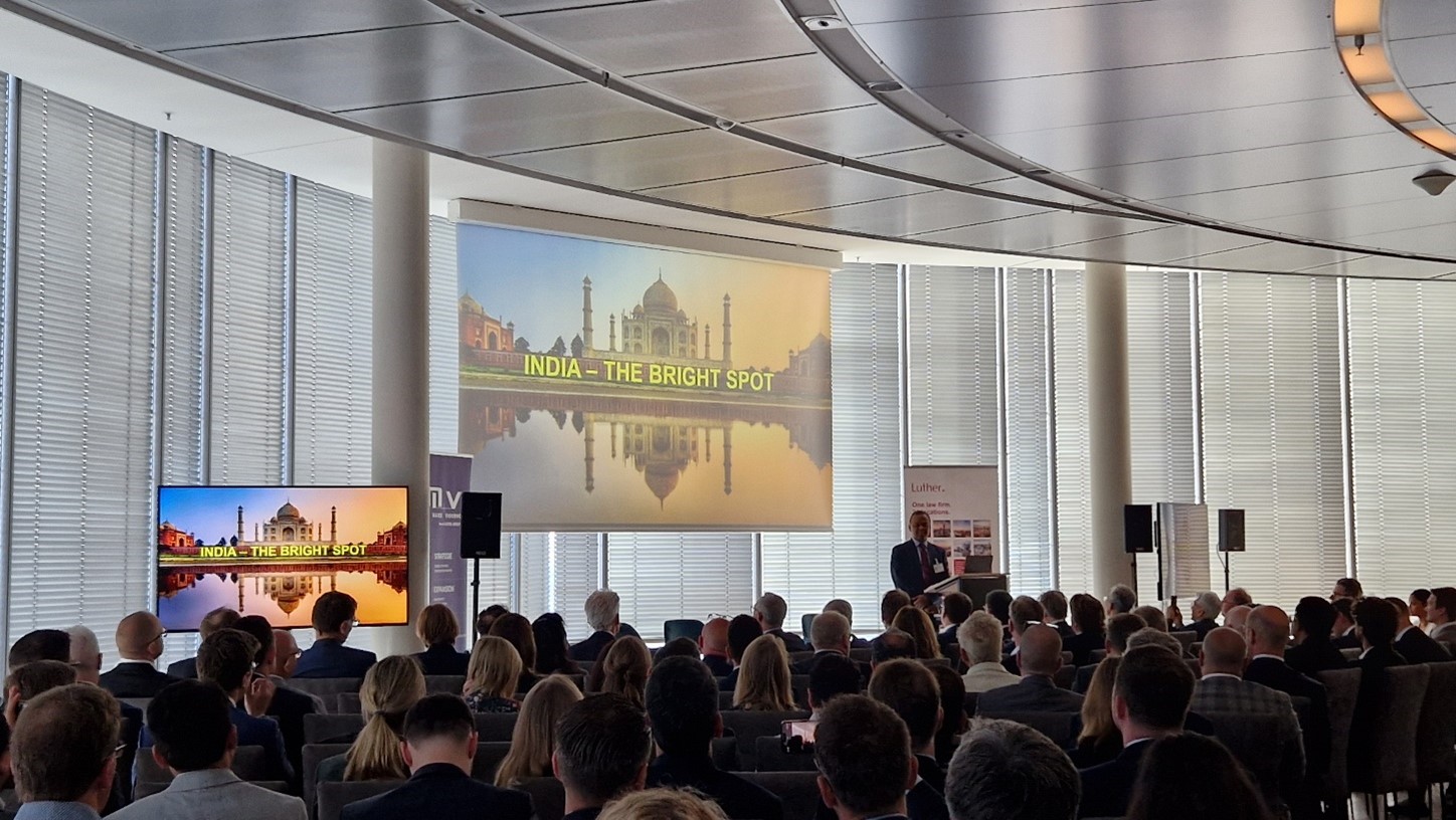 Ein Vortragender steht vor einem großen Bildschirm, auf dem die Worte „INDIA – THE BRIGHT SPOT“ über einem Bild des Taj Mahal und seines Spiegelbilds zu sehen sind. Im Vordergrund sitzen viele Zuschauer in einem modernen Veranstaltungsraum.