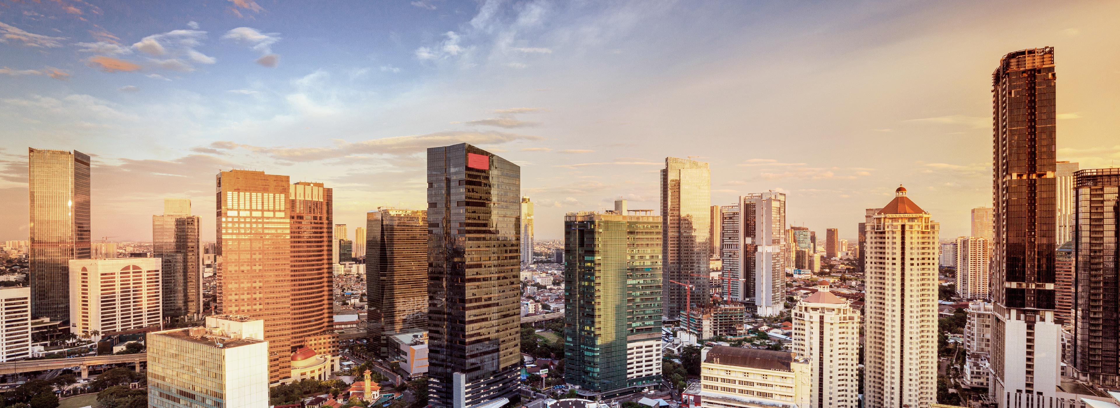 Eine Skyline mit modernen Hochhäusern bei Sonnenuntergang. Die Gebäude reflektieren warmes Licht, während der Himmel sanfte Farbtöne von Blau und Orange zeigt. Die Ansicht vermittelt den Eindruck einer lebhaften Stadtlandschaft.