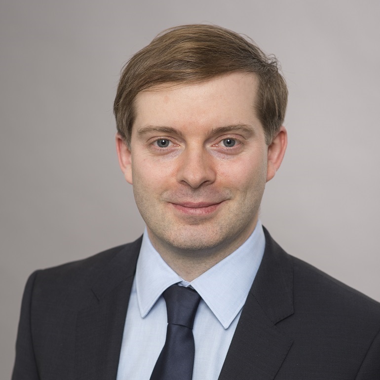 Eine junge, lächelnde Person mit kurzer, brauner Haarfrisur trägt einen dunklen Anzug und ein hellblaues Hemd. Der Hintergrund ist hell und neutral gehalten. Die Person schaut direkt in die Kamera.