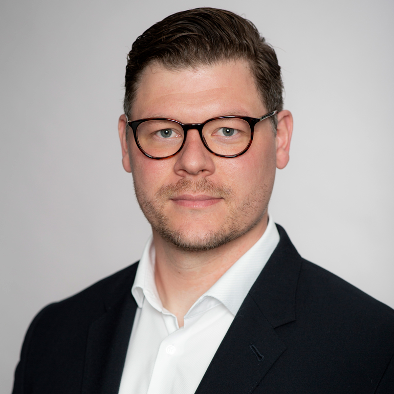 Ein Mann mit kurzen, dunklen Haaren und Brille steht vor einem einfarbigen, hellen Hintergrund. Er trägt einen schwarzen Blazer über einem weißen Hemd und blickt direkt in die Kamera. Sein Gesichtsausdruck ist neutral und freundlich.