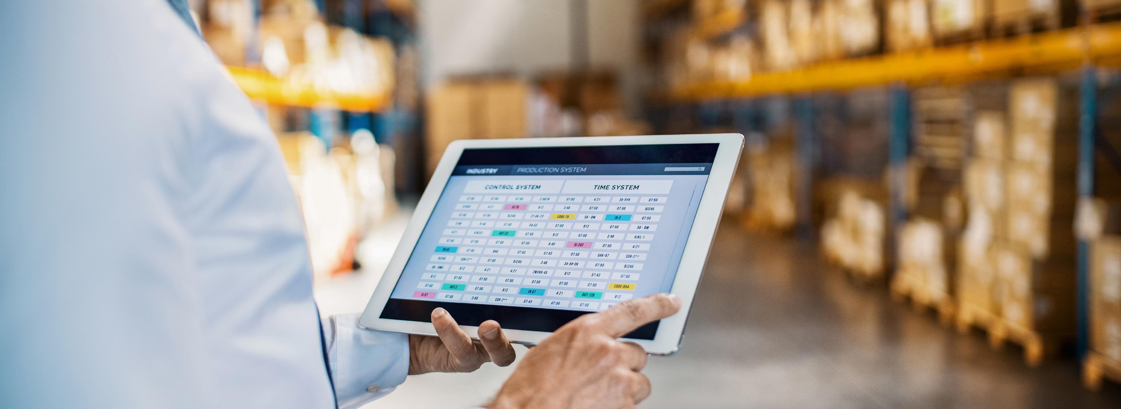 Eine Person hält ein Tablet in einer Lagerumgebung. Auf dem Bildschirm ist ein Kontrollsystem mit verschiedenen Daten und Zeitangaben zu sehen. Die Umgebung wirkt strukturiert, mit Regalen voller Kisten im Hintergrund, die den industriellen Kontext unterstreichen.