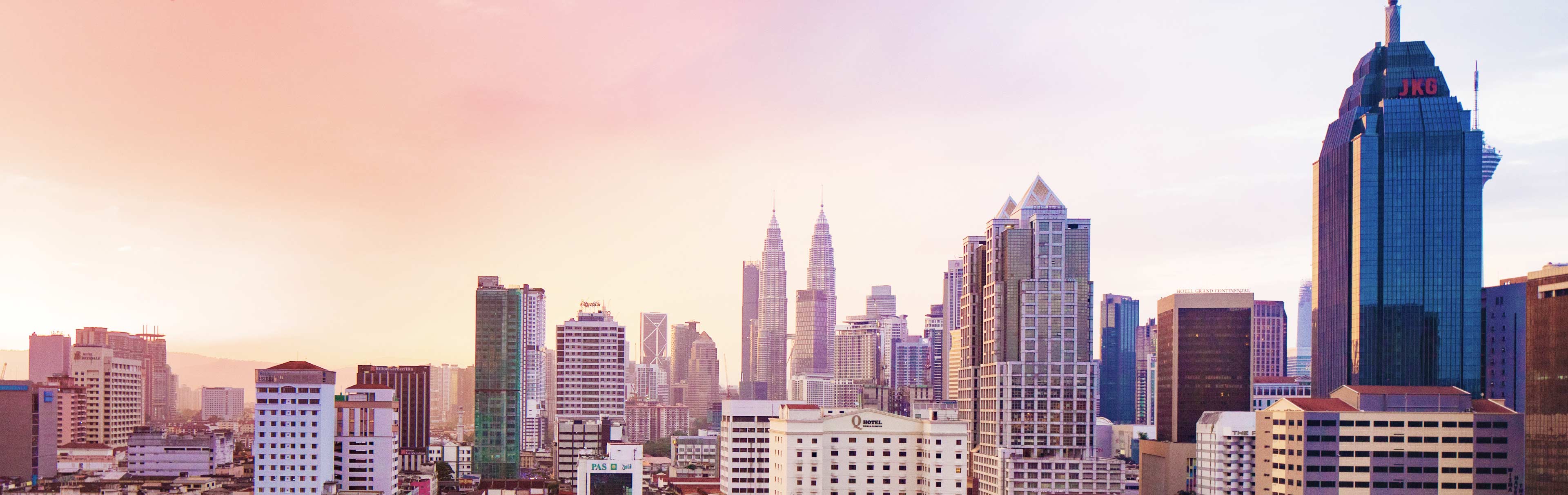 Der Blick auf die Skyline von Kuala Lumpur zeigt moderne Wolkenkratzer, darunter die markanten Petronas Twin Towers. Der Himmel ist in sanften Rosa- und Lilatönen gefärbt, was eine ruhige Abendstimmung erzeugt. Die Architektur vereint unterschiedliche Stile und spiegelt die Dynamik der Stadt wider.