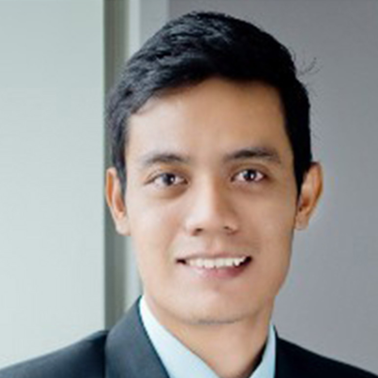 Porträt eines jungen Mannes in Anzug und Krawatte, lächelnd. Er hat dunkles, kurzes Haar und trägt ein hellblaues Hemd unter seinem Anzug. Der Hintergrund ist neutral und unscharf, wodurch der Fokus auf dem Gesicht des Mannes liegt.