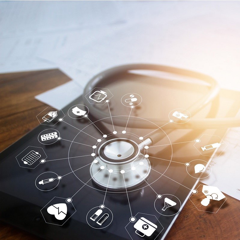 A stethoscope is positioned on a digital tablet, surrounded by icons representing various healthcare tools and services, such as medical equipment, prescriptions, and healthcare symbols. The background features scattered papers, emphasizing a medical or clinical setting.