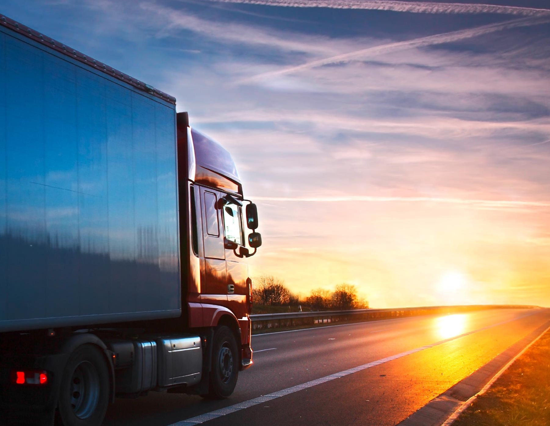 A large red truck drives along an empty highway at sunset. The sky is filled with soft hues of orange and pink, casting a warm glow on the road. The truck's silhouette is prominent against the colorful backdrop, emphasizing a serene travel scene.