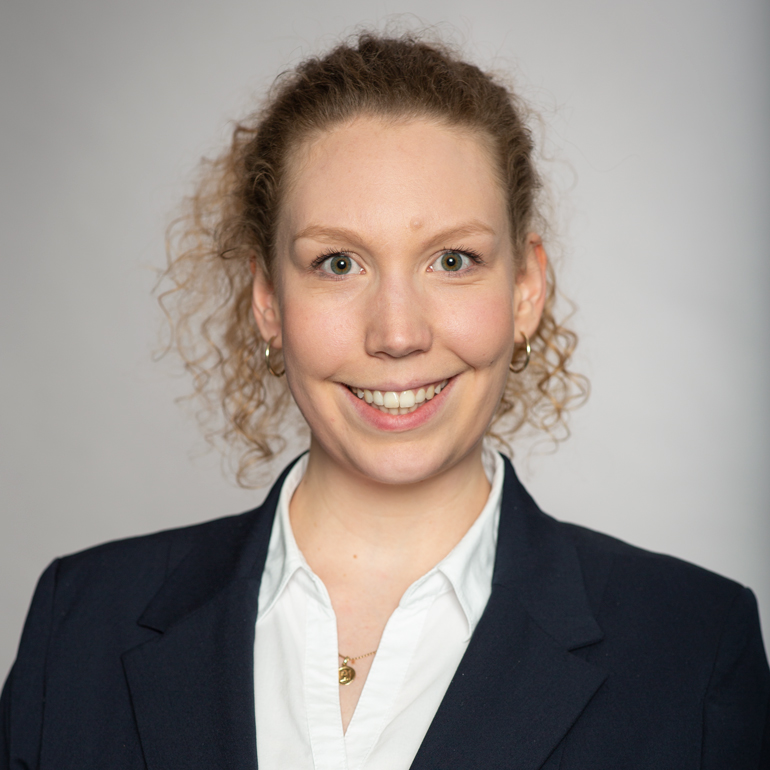 Porträt einer lächelnden Frau mit lockigem, blonden Haar, die einen dunkelblauen Blazer und ein weißes Hemd trägt. Der Hintergrund ist hell und neutral. Die Frau sieht freundlich und selbstbewusst aus.