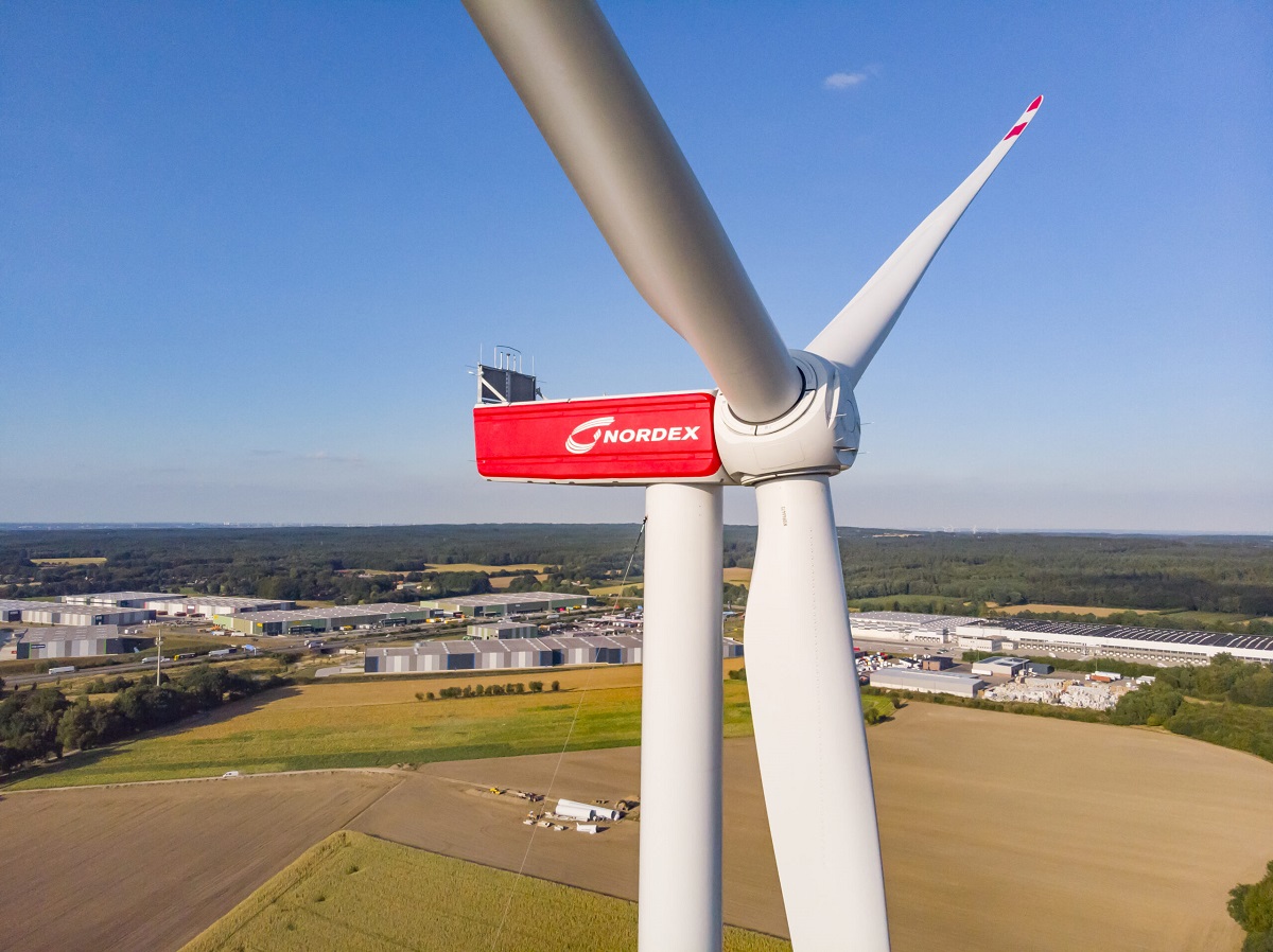 A close-up view of a red Nordex wind turbine, showcasing its large blades against a clear blue sky. The background features fields and industrial buildings, highlighting a renewable energy landscape.