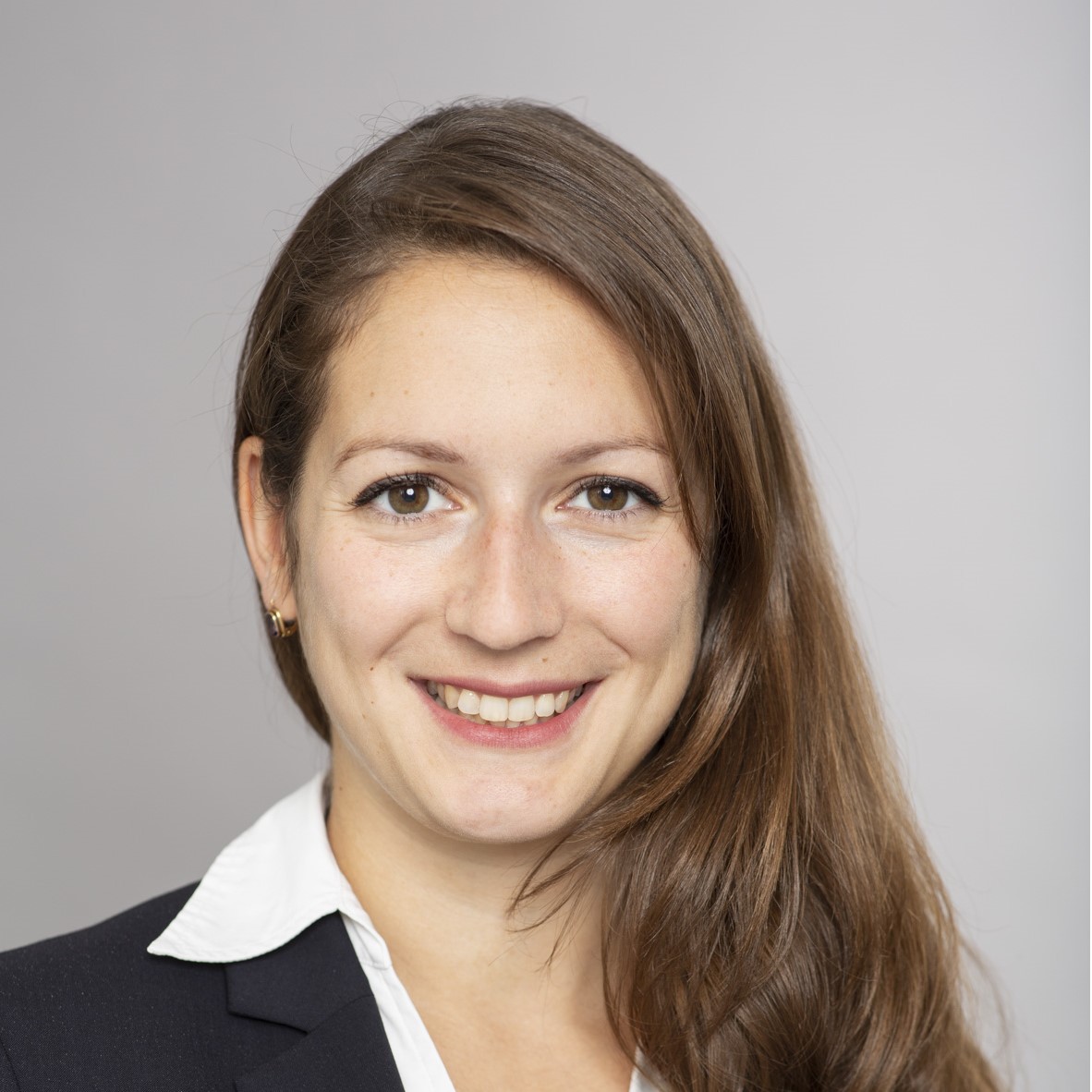 Porträt einer lächelnden Frau mit langen, braunen Haaren, die einen schwarzen Blazer und ein weißes Hemd trägt. Der Hintergrund ist hell und einfarbig. Die Frau hat einen freundlichen Ausdruck.