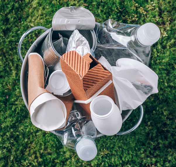 Ein Mülleimer ist mit verschiedenen Abfallarten gefüllt, darunter Plastikflaschen, Papiertüten, Aluminiumdosen und Verpackungsmaterial. Der Eimer steht auf grünem Gras.