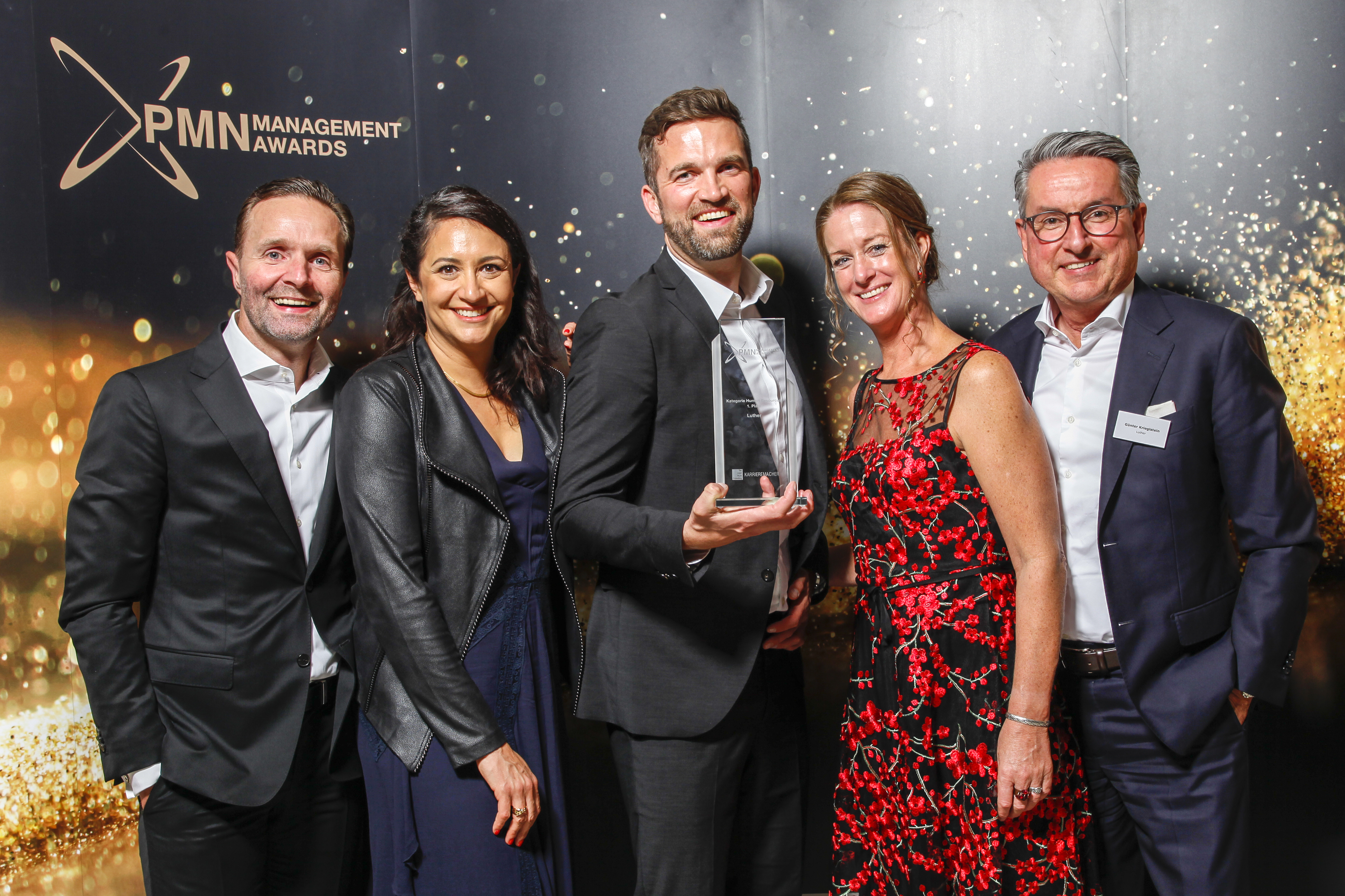 Fünf Personen stehen zusammen und lächeln für ein Foto bei den PMN Management Awards. Eine Person hält einen Preis in der Hand. Die Umgebung ist festlich beleuchtet, mit goldenen Akzenten im Hintergrund.