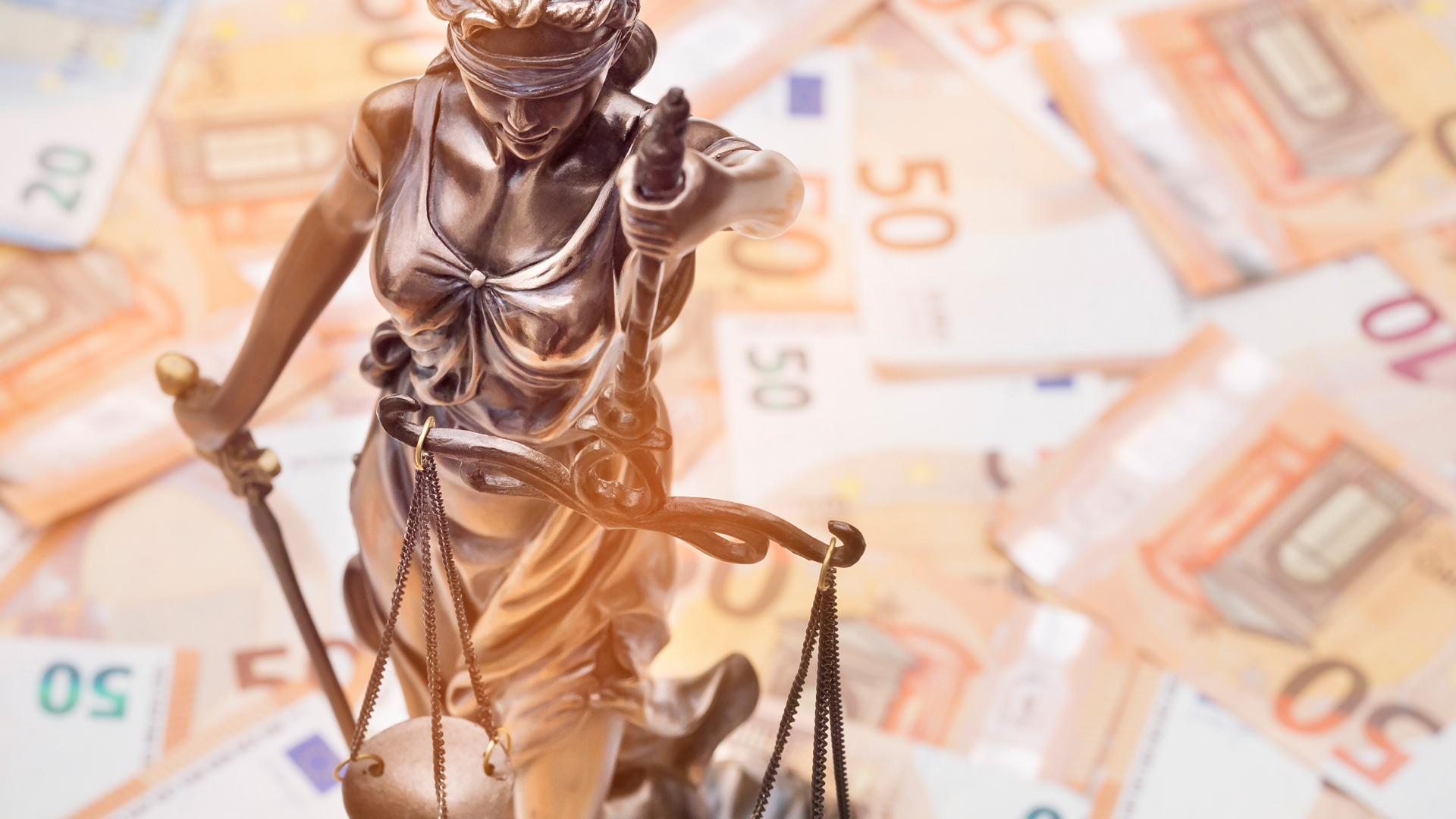 Eine Skulptur der Justitia, die eine Waage in der einen und ein Schwert in der anderen Hand hält. Im Hintergrund liegen zahlreiche Geldscheine. Die Darstellung symbolisiert Gerechtigkeit und die Verbindung von Recht und Geld.