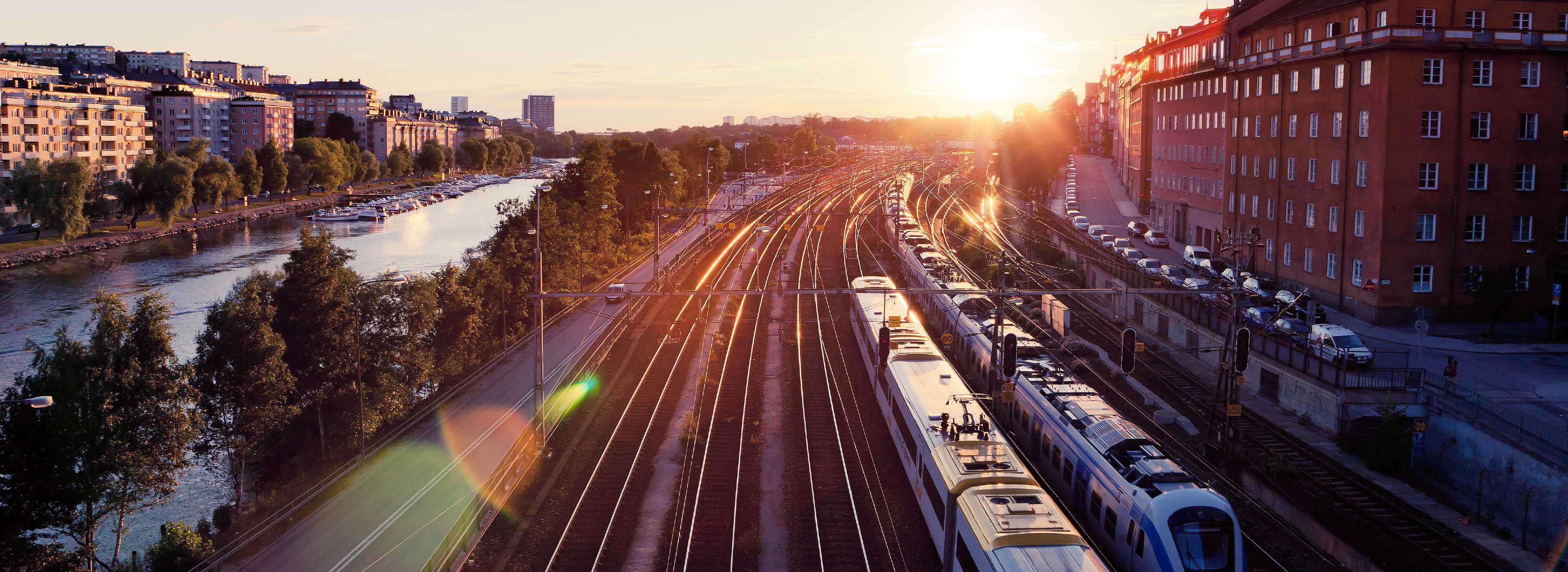 Ein Sonnenuntergang über einem Bahnhof mit Gleisen, die parallel verlaufen. Auf der linken Seite fließt ein Fluss, umgeben von Bäumen. Entlang des Ufers stehen Boote. Im Hintergrund sind Wohngebäude sichtbar. Die Szenerie strahlt eine ruhige, stimmungsvolle Atmosphäre aus.
