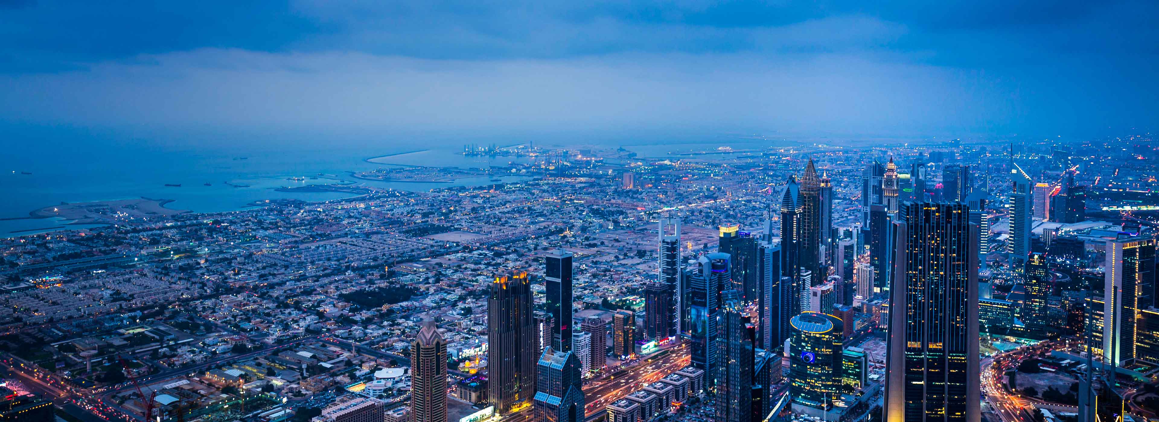 Eine Panoramaansicht einer modernen Stadt bei Dämmerung. Hohe Wolkenkratzer erleuchten die Umgebung mit zahlreichen Lichtern. Im Hintergrund ist das Meer sichtbar, ergänzt durch einige Schiffe. Die Atmosphäre ist ruhig und zeitgenössisch, mit einem klaren urbanen Charme.