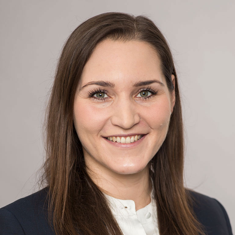 Portrait einer Frau mit langen, braunen Haaren und freundlichem Lächeln. Sie trägt eine dunkelblaue Blazerjacke und ein helles Oberteil. Der Hintergrund ist neutral und unauffällig. Die Frau strahlt Selbstbewusstsein und Freundlichkeit aus.