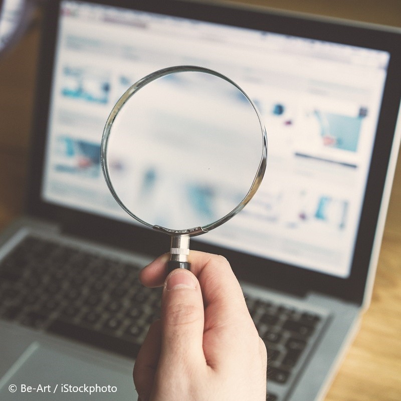 A hand holding a magnifying glass is positioned in front of a laptop screen displaying various web content. The image symbolizes attention to detail or searching for information.