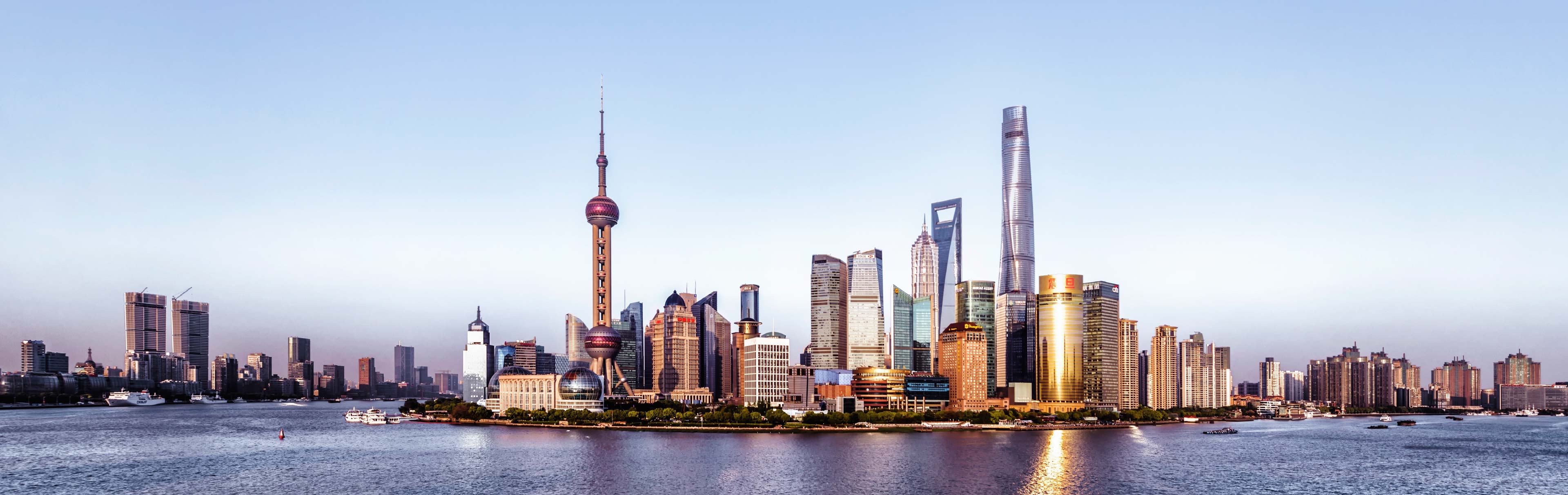 Ein Panoramablick auf die Skyline von Shanghai bei Sonnenaufgang. Hohe Wolkenkratzer und moderne Gebäude dominieren das Stadtbild, darunter der markante Oriental Pearl Tower. Der Huangpu-Fluss spiegelt das Licht der Stadt wider und verleiht der Szene eine beeindruckende Atmosphäre.