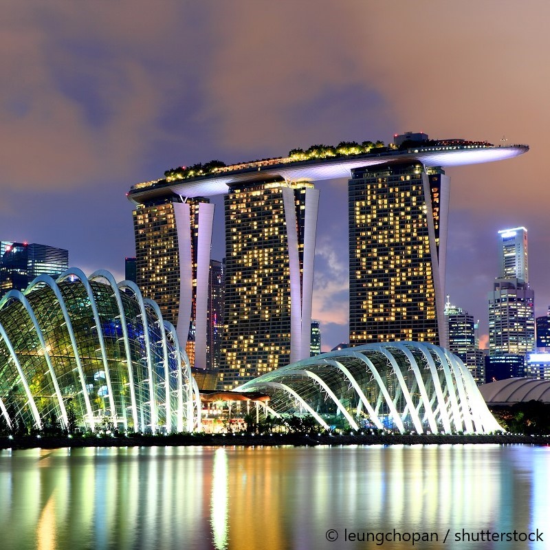 A stunning skyline featuring the iconic Marina Bay Sands hotel with its elevated garden rooftop, illuminated against a twilight sky. In the foreground, the futuristic design of the Gardens by the Bay showcases its glass structures, reflecting beautifully in the calm water below.