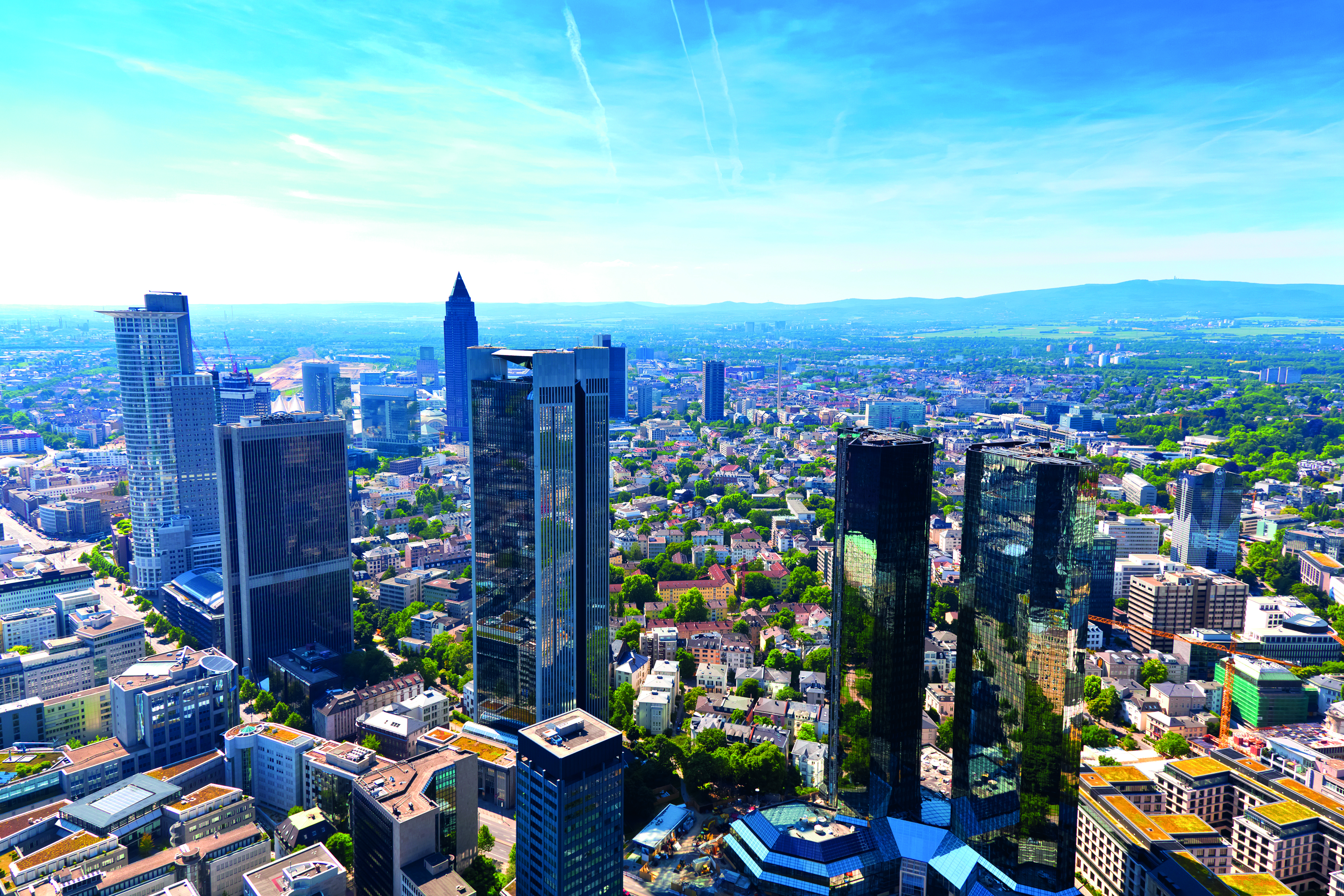 A panoramic view of a bustling cityscape featuring several tall skyscrapers and modern buildings. Lush green areas and residential neighborhoods are visible among the urban landscape, under a clear blue sky with few clouds, showcasing a vibrant city environment.
